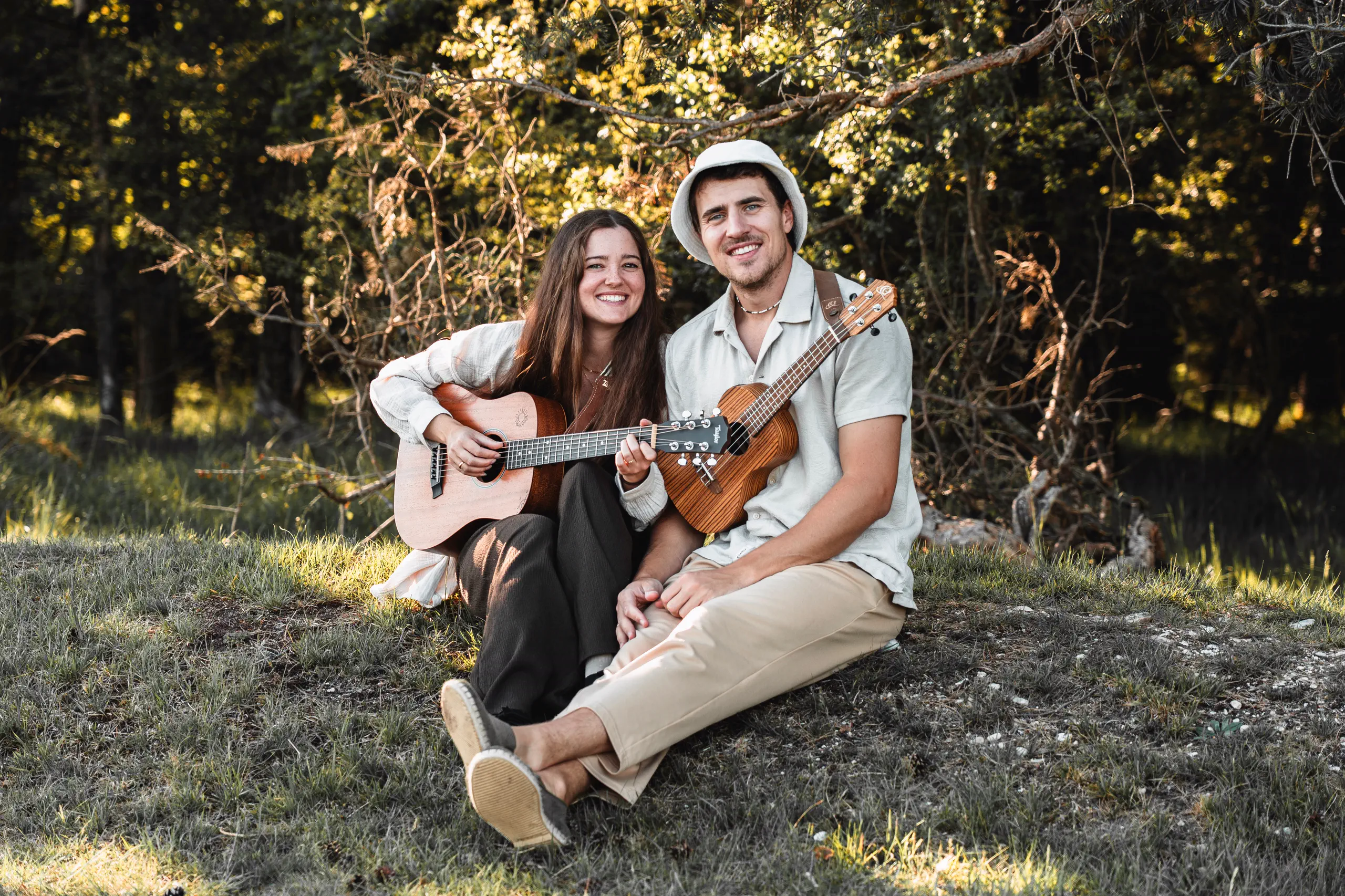 Sunny und Marty sitzen mit Gitarre und Ukulele im Gras unter Bäumen und lachen in die Kamera